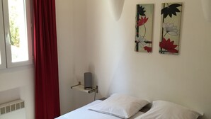 A neatly made bed with white linens, a red-striped blanket, and two pillows. There are two wall-mounted light fixtures and two floral wall art pieces. A window with a red curtain is visible, and a small heater is mounted on the wall. - Villa STELLA D ORO, F2 * (Porto-Vecchio)