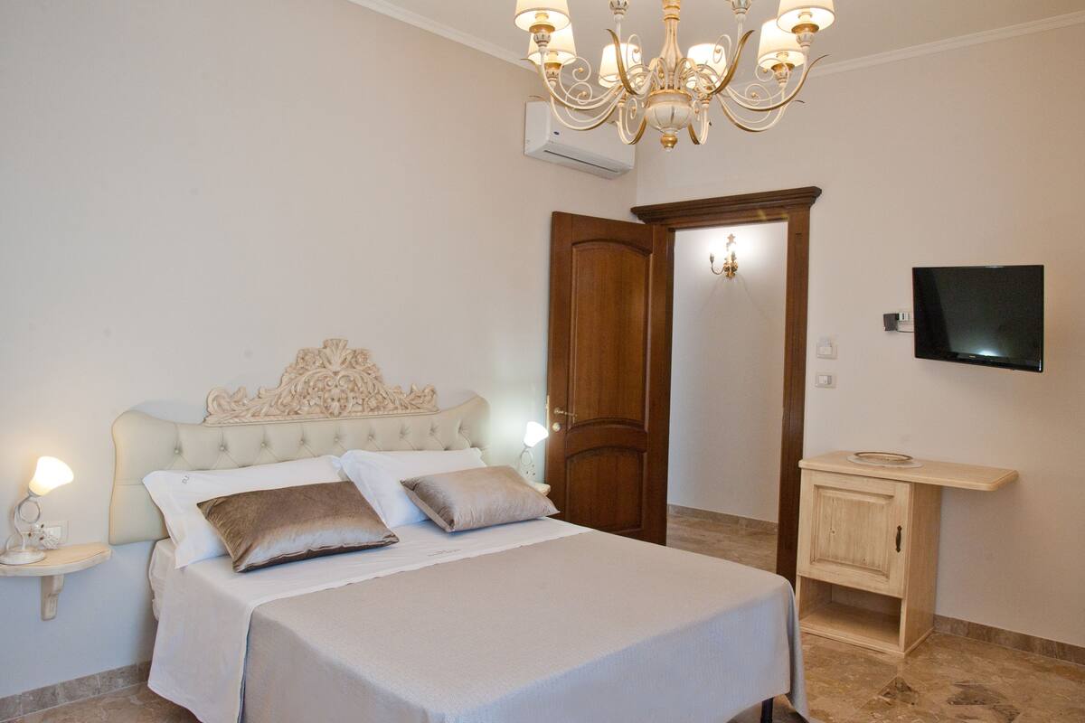 A bedroom with a bed, bedside table, lamp, chandelier, TV, and a wooden door.