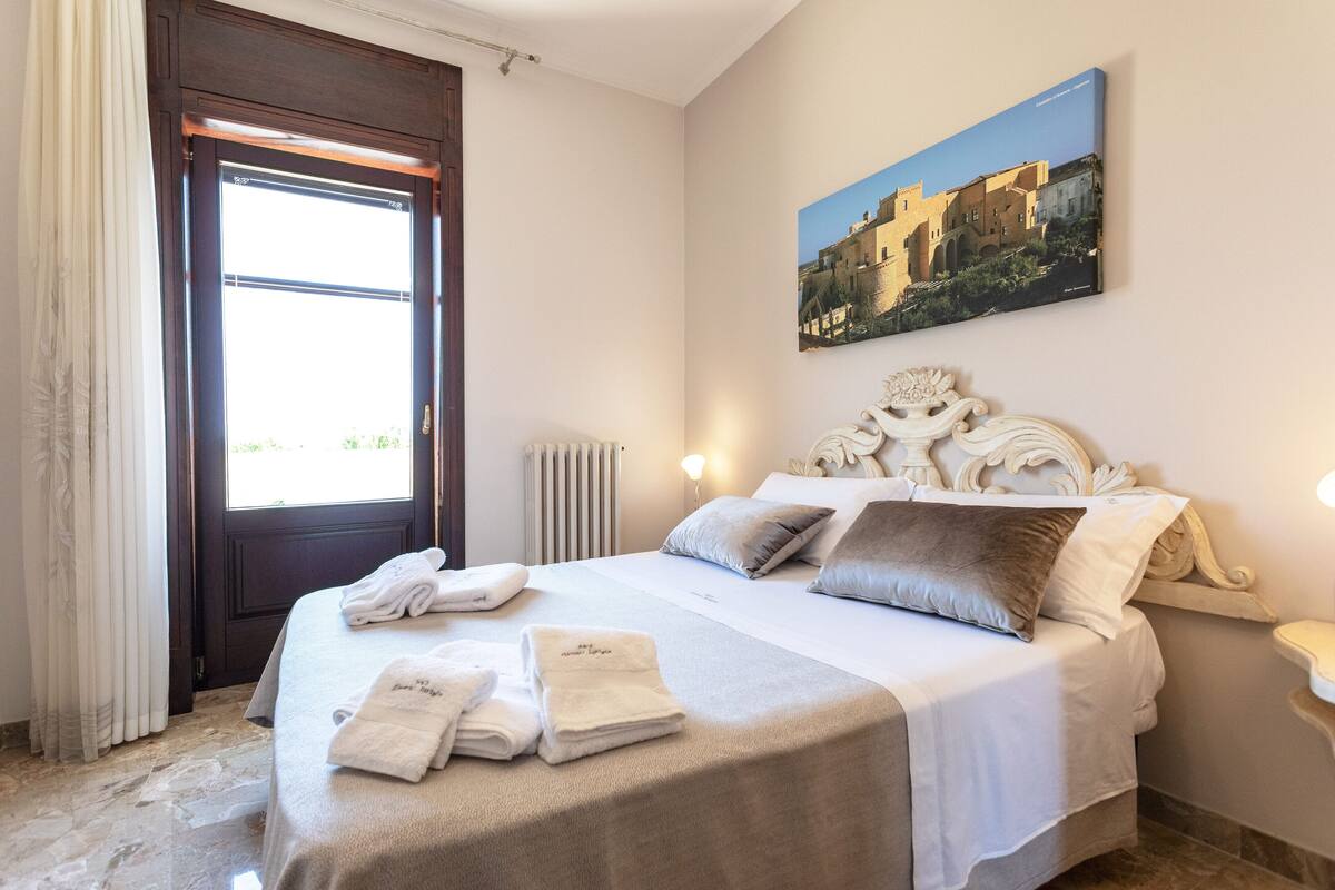 A neatly made bed with towels, a painting of a building on the wall, and a window with curtains.
