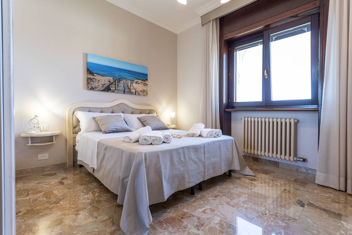 A bedroom with a bed, a bedside table, a painting, a window, and a radiator.