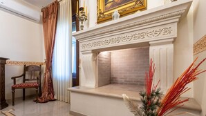 A room with a fireplace, a chair, a vase with plants, and a window with curtains. - Domus Japigia (Ugento)