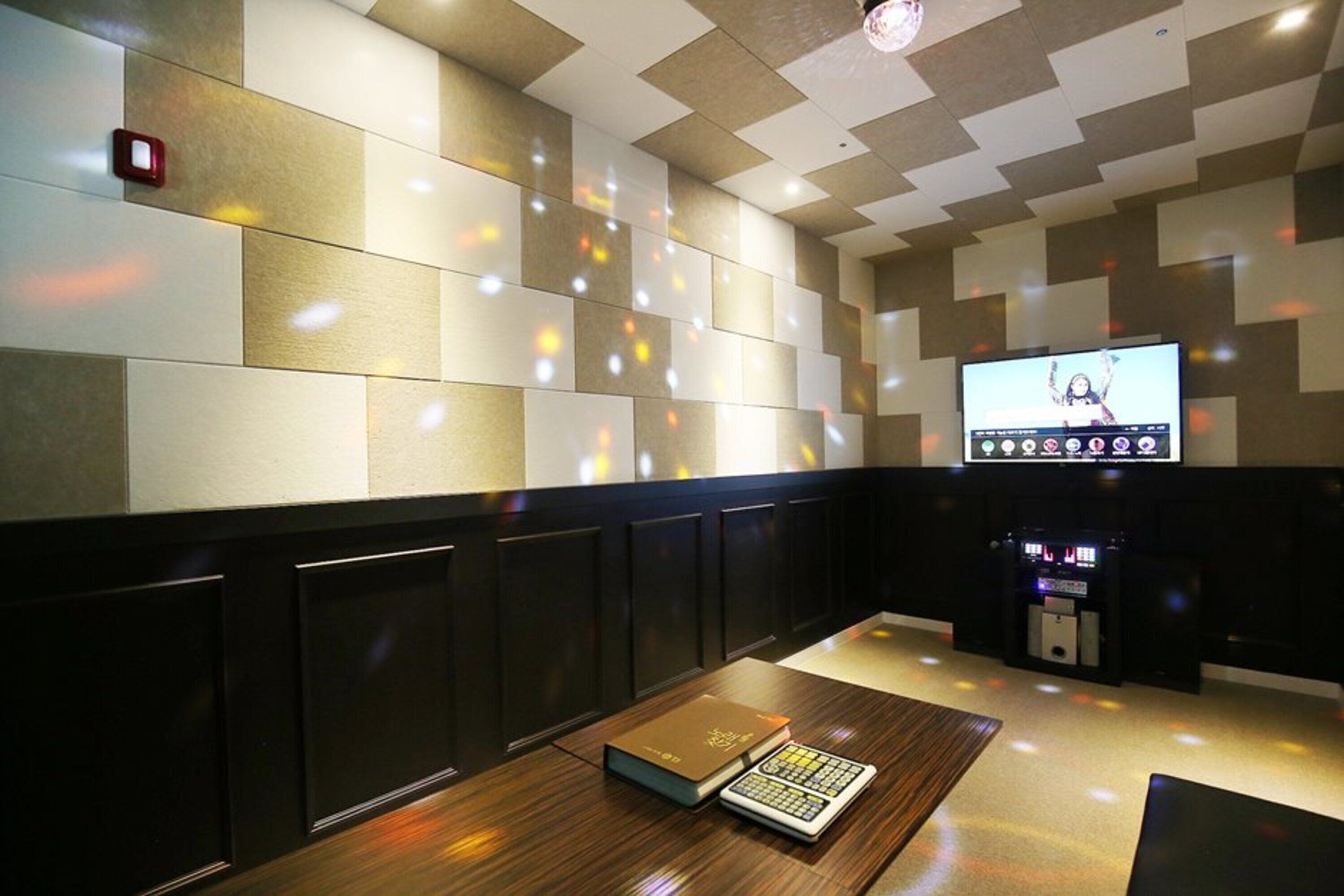 A modern room with a checkered wall pattern, a flat-screen TV, a console table with a book and a calculator, and a ceiling with recessed lighting.