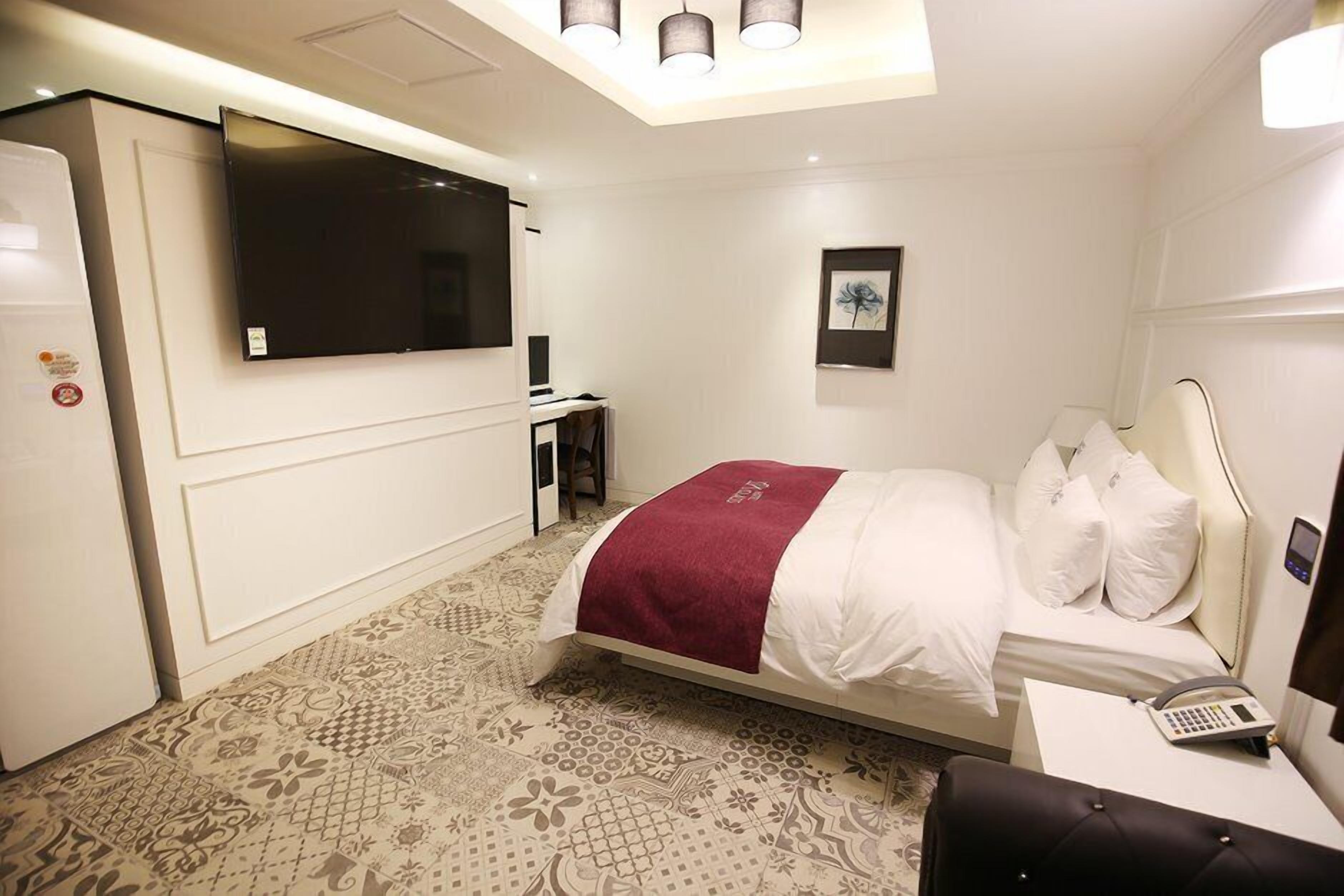 A hotel room with a bed, a desk with a telephone, a television mounted on the wall, and a patterned carpet.