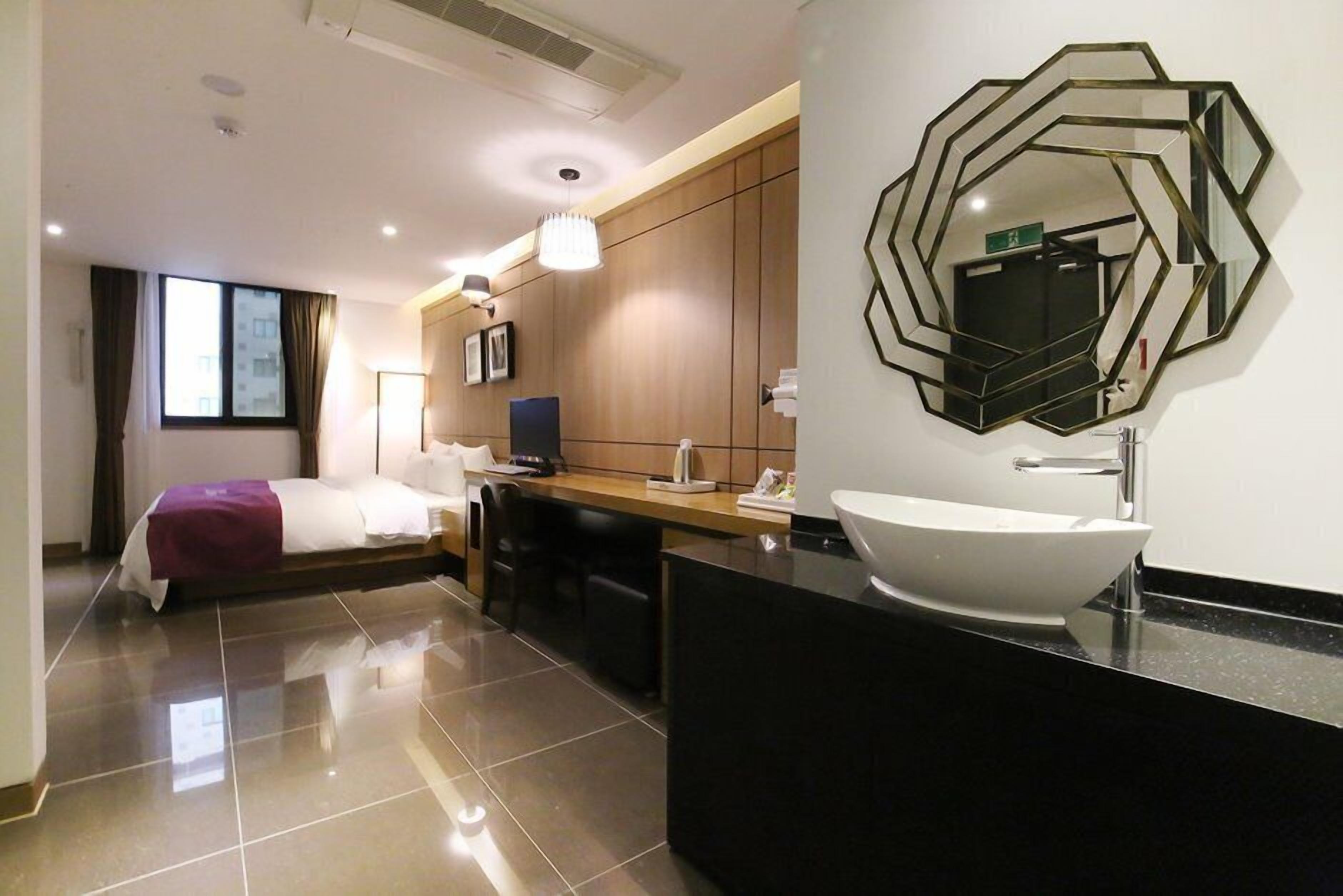 A modern hotel room with a large bed, a desk, a bathroom with a large mirror, and a black countertop.