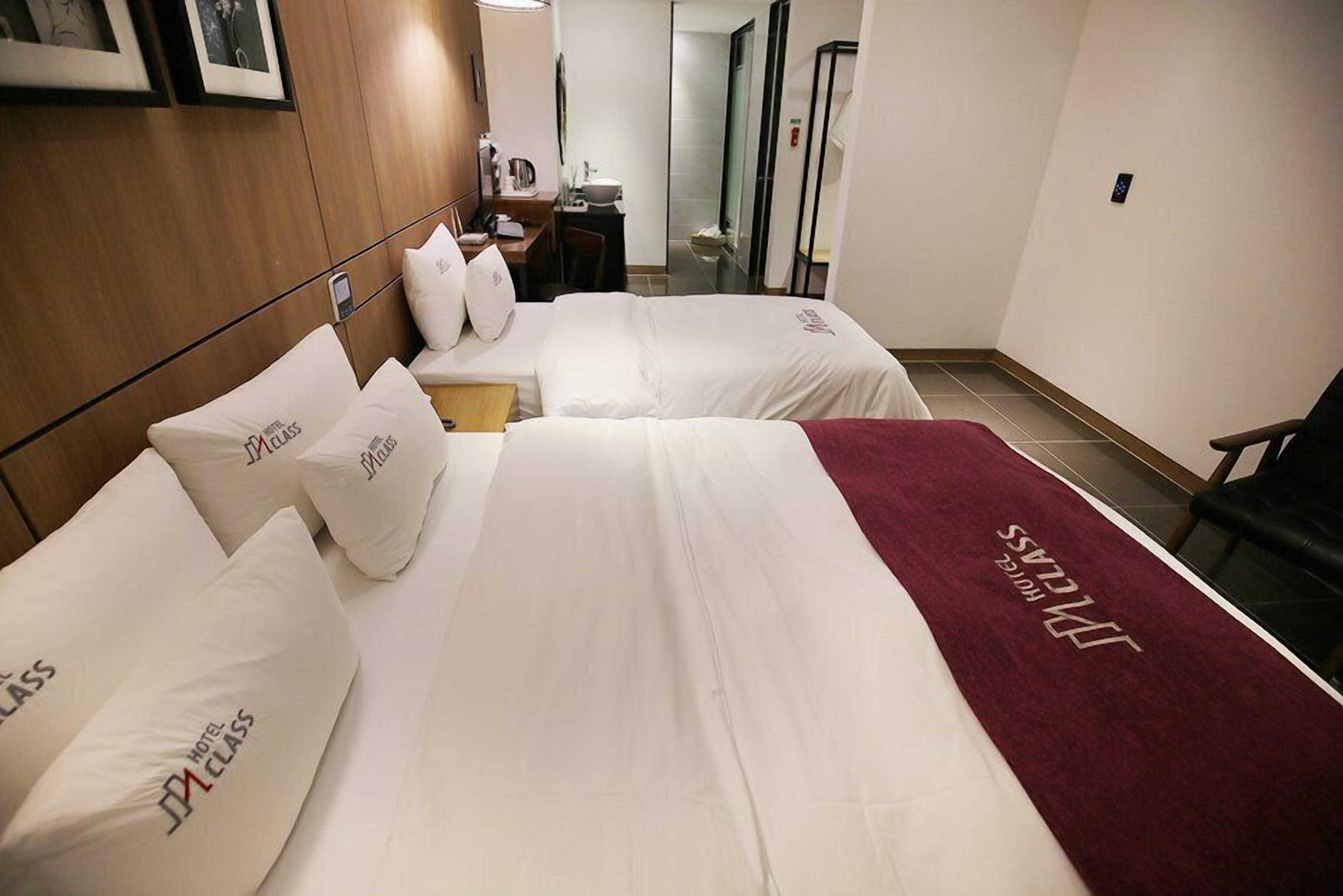 A hotel room with two beds, each with a pillow labeled "HOTEL CLASS." There is a wooden headboard, a desk with a chair, and a mirror.