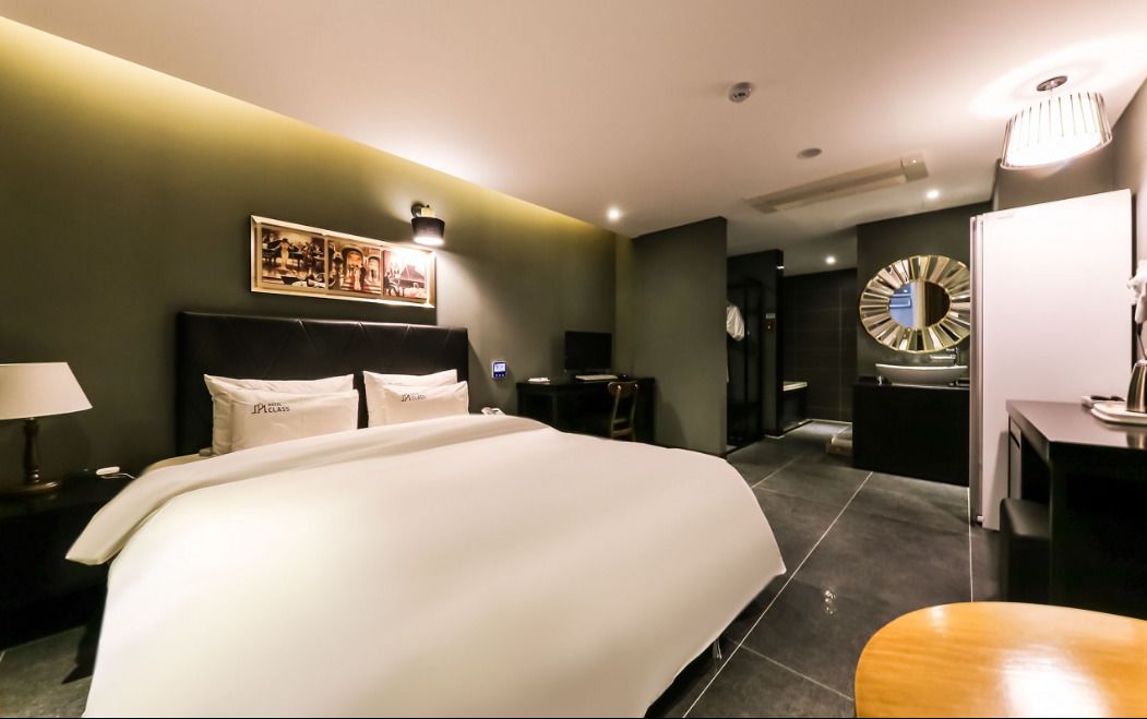 A modern hotel room with a large bed, a desk, a vanity with a mirror, and a small seating area.