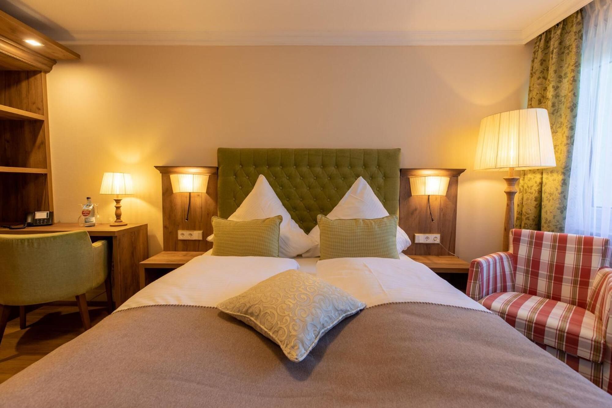 Une chambre d’hôtel avec un grand lit, deux tables de chevet avec des lampes, une tête de lit recouverte d’un tissu vert et un fauteuil à motifs écossais.