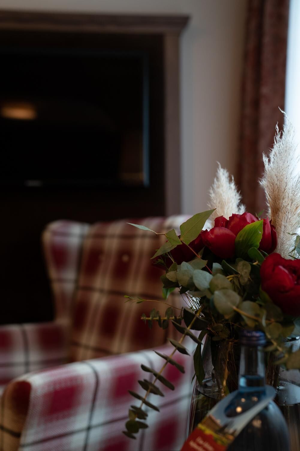 Una habitación con un sofá a cuadros, un jarrón de rosas rojas y hierba de pampa.