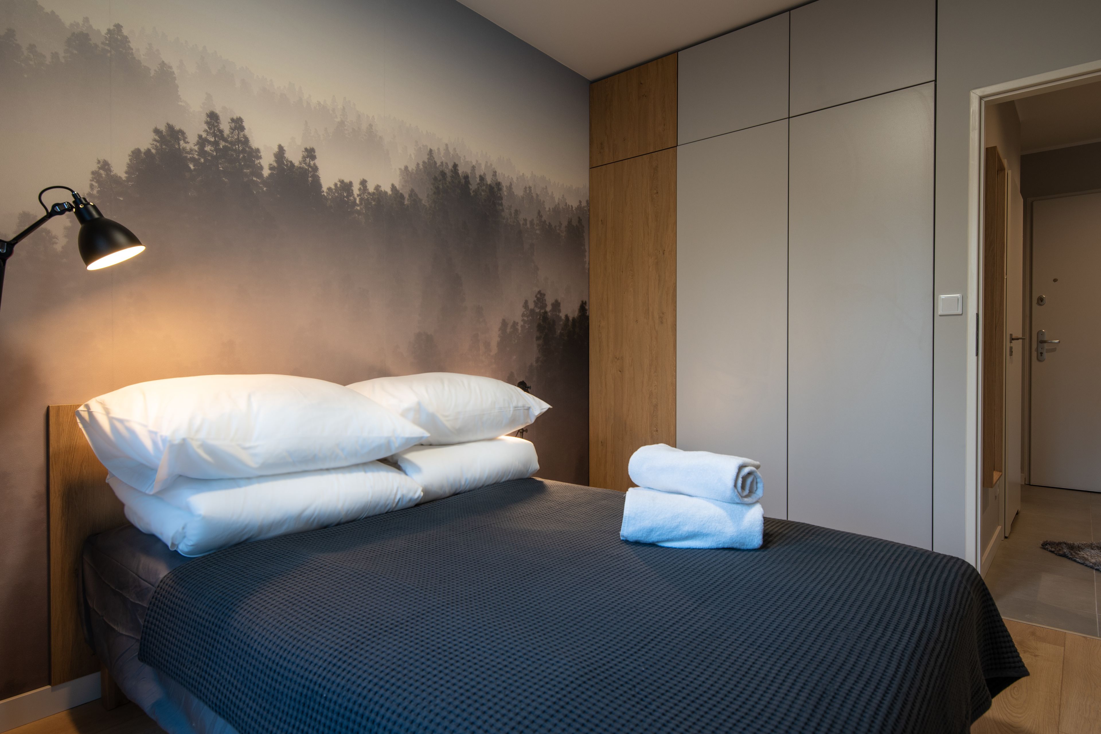 Cama bem arrumada com travesseiros brancos e toalha dobrada, luminária de cabeceira e painel na parede com desenho de uma floresta.