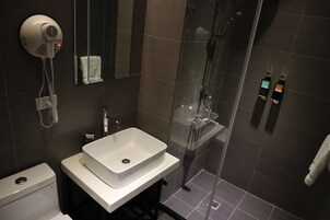 Un baño moderno con un lavamanos blanco, un secador de pelo montado en la pared, una ducha con cerramiento de vidrio y dos dispensadores de jabón en la pared.