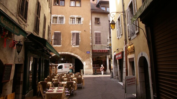 A narrow street with tables and chairs set up for outdoor dining, flanked by buildings with various signs and a van parked in front. - Studio 2 people with parking Annecy (Annecy)
