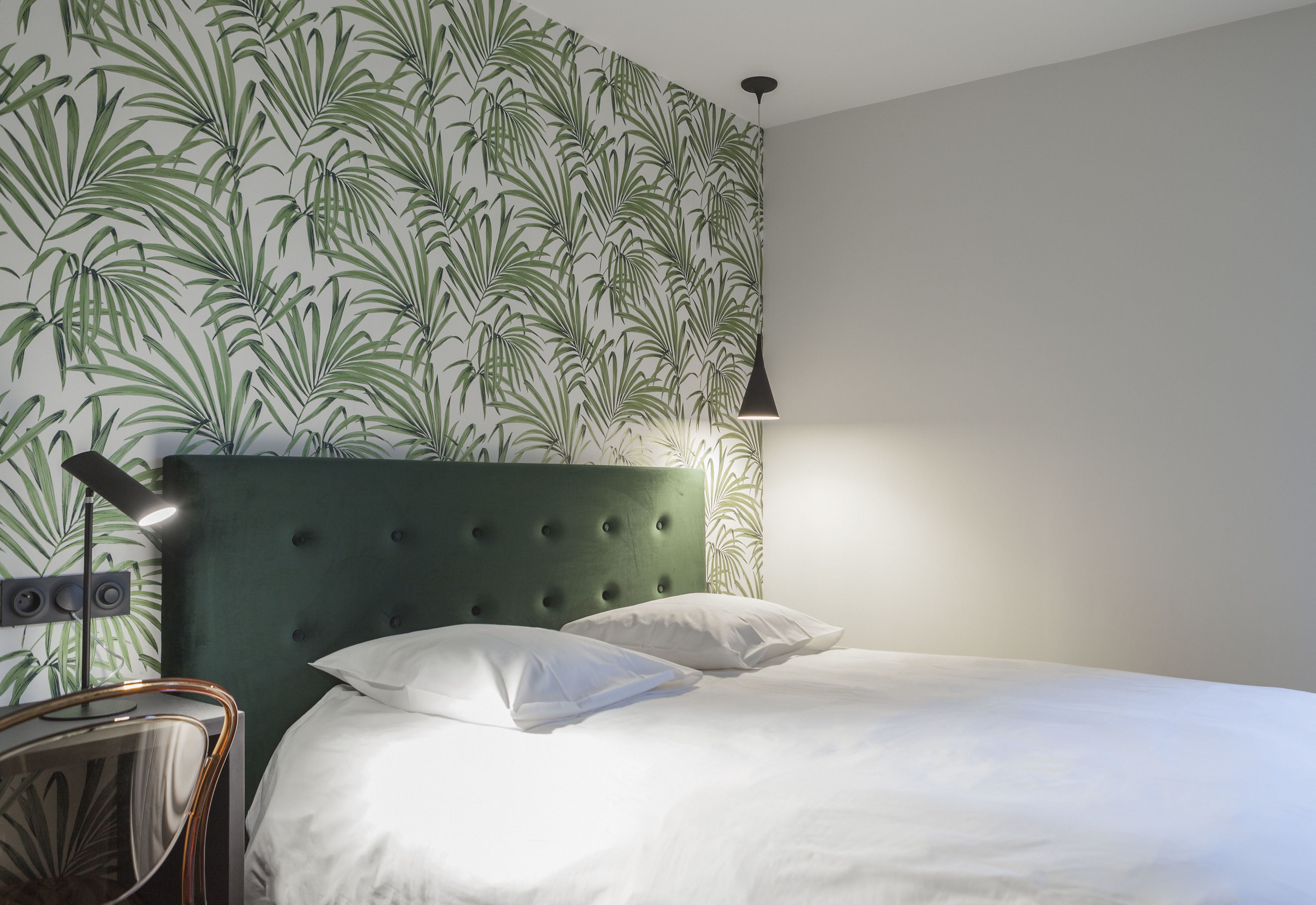 A neatly made bed with white linens, a green upholstered headboard, and a wall with a leafy pattern wallpaper.