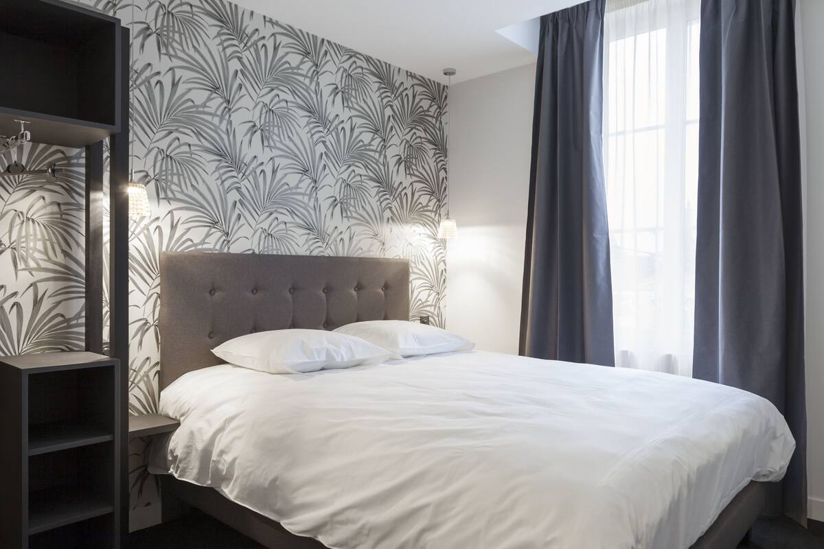 A neatly made bed with white linens, a tufted headboard, and a wall with a leafy pattern wallpaper.