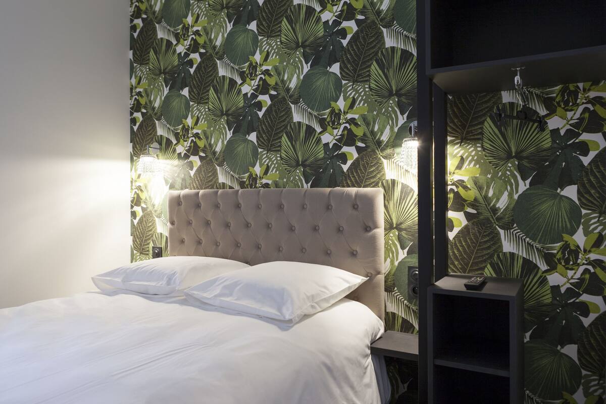 A bedroom with a tufted headboard, white bedding, and a wall with a tropical leaf pattern.