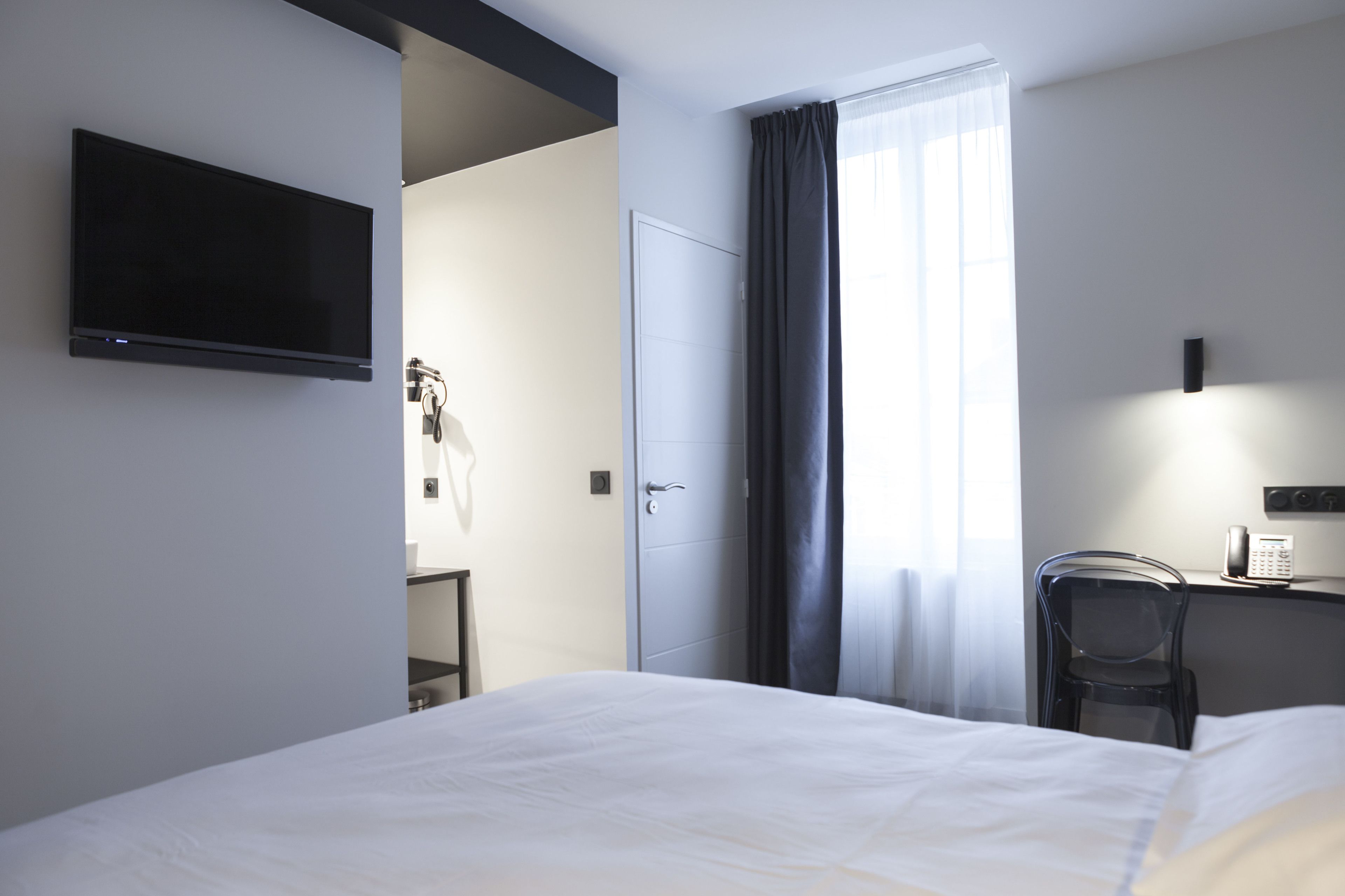 A hotel room with a bed, a desk with a chair, a TV mounted on the wall, and a door leading to a bathroom.