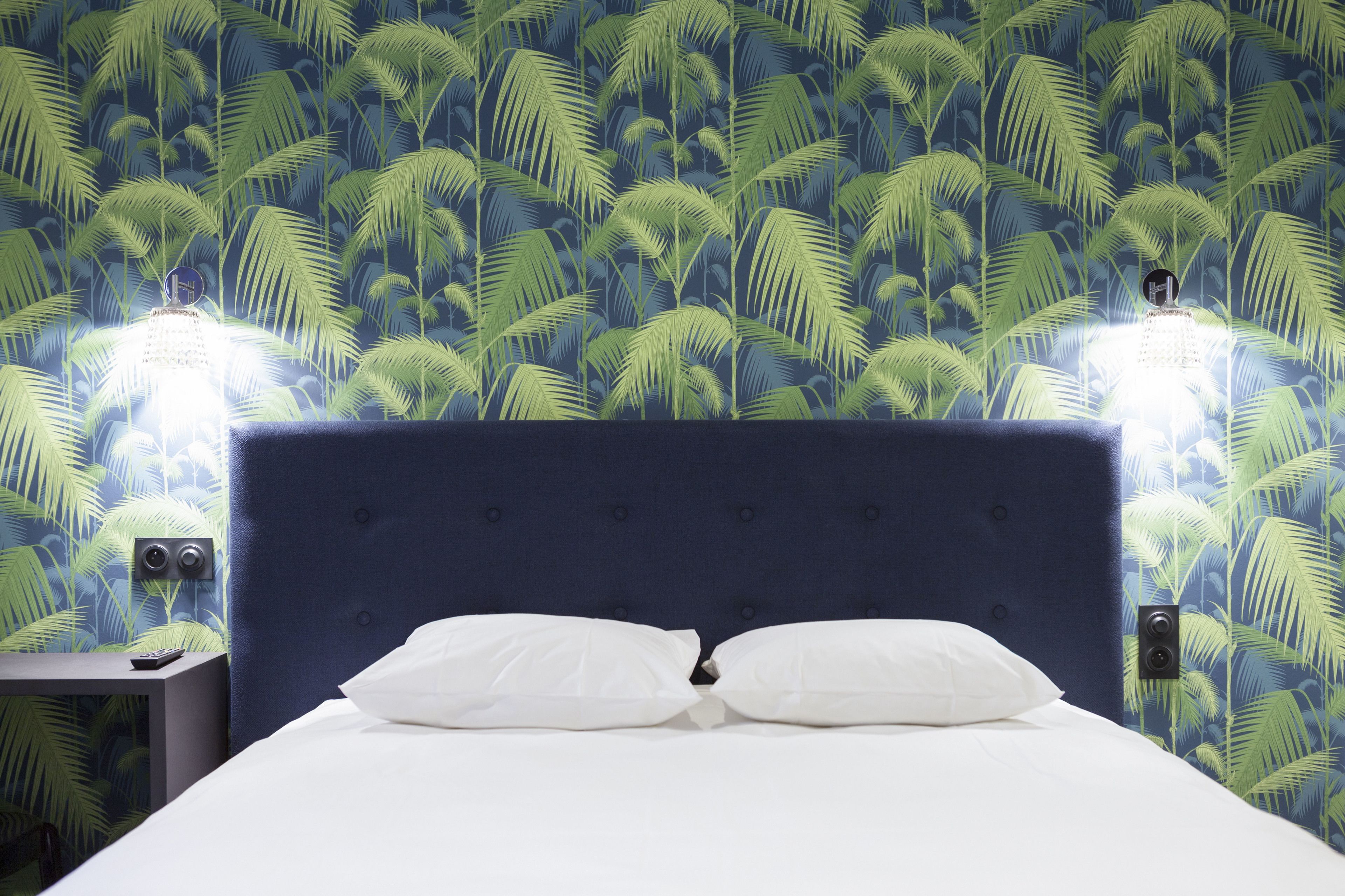 Une chambre d’hôtel avec un lit, des oreillers blancs, une tête de lit bleu foncé et un mur orné d’un motif de feuilles de palmier.