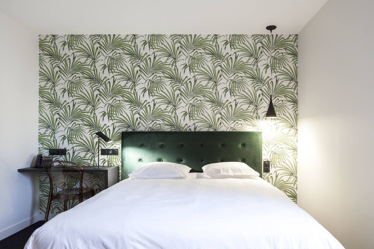 A neatly made bed with white linens, a green upholstered headboard, and a wall with a leafy pattern wallpaper.