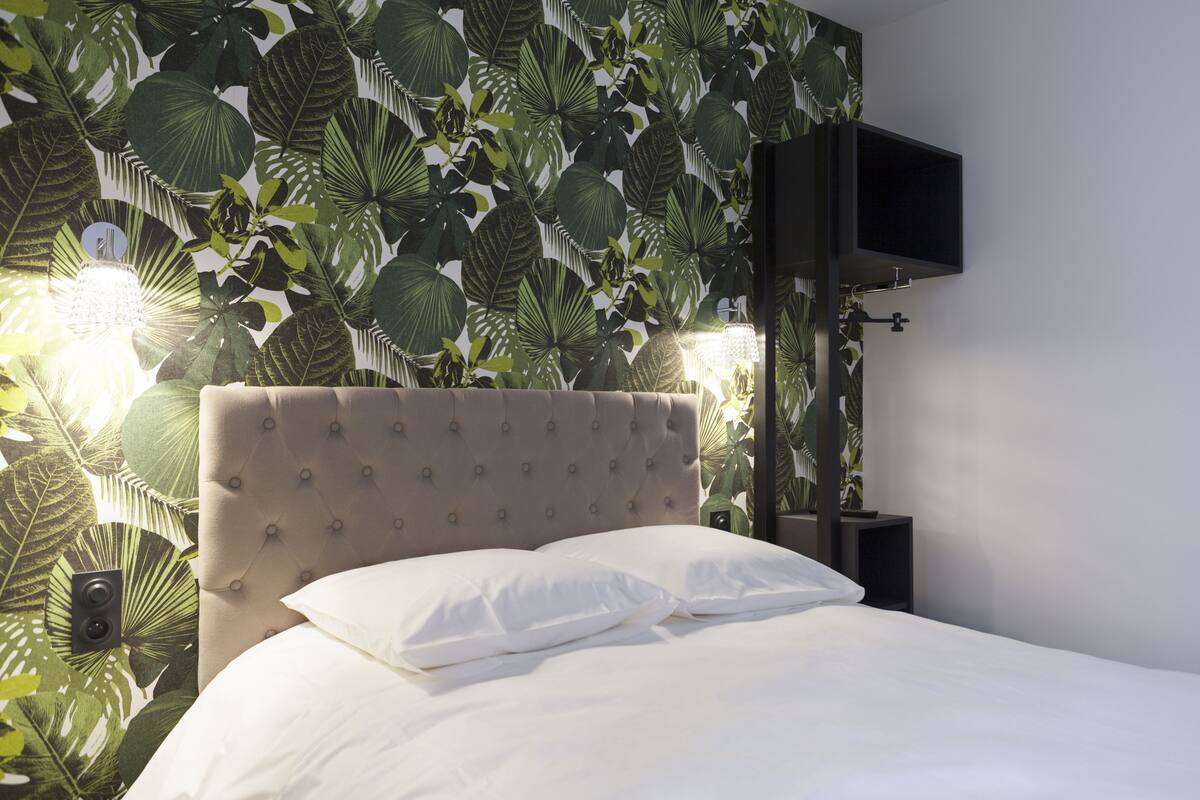 A bedroom with a tufted headboard, a bed with white linens, a wall-mounted TV, and a tropical leaf-pattern wallpaper.