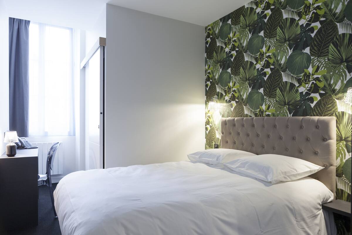 A hotel room with a bed, a desk with a lamp, a chair, a window with curtains, and a wall with a tropical leaf pattern.