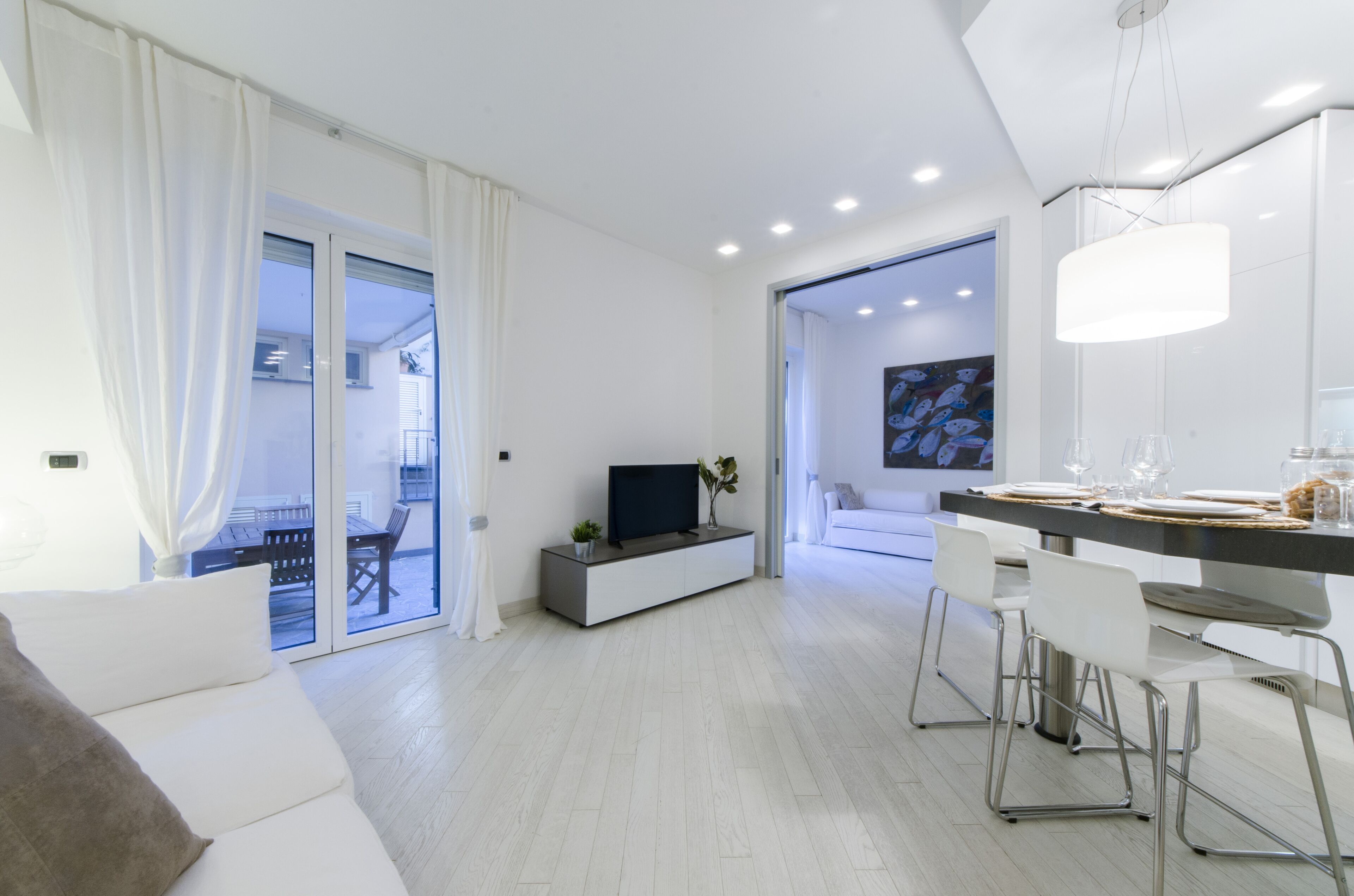 A modern living room with a sofa, a TV on a low white cabinet, a dining area with a table and chairs, and a large window with curtains.