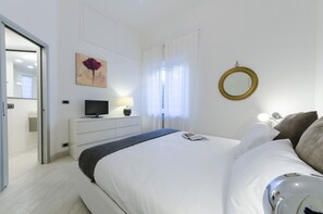 A modern hotel room with a large bed, a flat-screen TV, a bedside table with a lamp, a wall-mounted mirror, and a door leading to a bathroom.