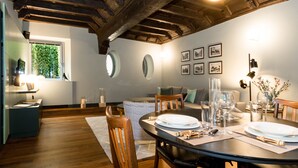 Un coin repas avec une table ronde dressée pour quatre personnes, entourée de chaises en bois. Des tableaux encadrés ornent le mur, une fenêtre offre une vue sur la verdure et le plafond est en bois.