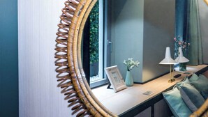 Une pièce avec un grand miroir, une fenêtre donnant sur un paysage verdoyant, une table de chevet avec une lampe, un vase de fleurs et un tableau encadré.
