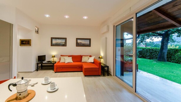 A modern living room with a red sofa, a coffee table with a teapot, and a large sliding glass door leading to a garden. - JOIVY Golf Garlenda - Charming Villa Guest House (Garlenda)