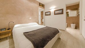 A bedroom with a bed, bedside table, lamp, and a bathroom visible through an open door. - JOIVY Golf Garlenda - Charming Villa Guest House (Garlenda)