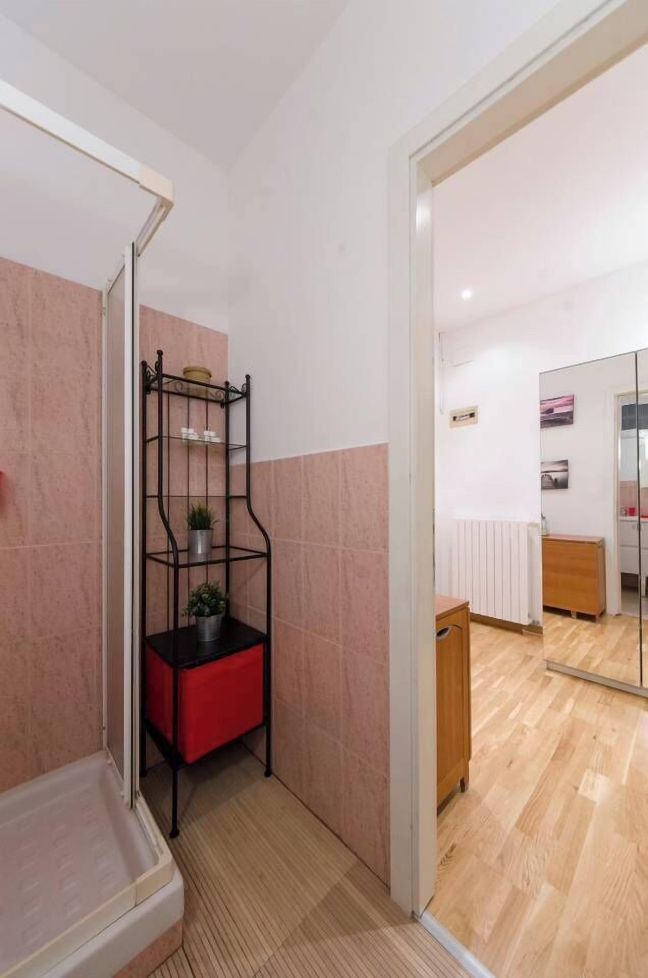 A bathroom with a shower, a black metal shelf with plants, a red chair, and a wooden cabinet.