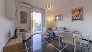 A modern living room with a sofa, a dining table set for two, a TV, and a large window with a view of the city. - JOIVY Family Flat Vicino al Mare a Genova Quinto (Genoa)