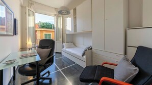A compact room with a bed, a desk, and a chair. There is a window with curtains, a lamp, and a view of buildings outside. - JOIVY Family Flat Vicino al Mare a Genova Quinto (Genoa)