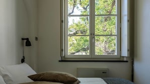 Une chambre avec une grande fenêtre, un lit avec une literie blanche et marron, une lampe de bureau noire et un radiateur placé sous la fenêtre.