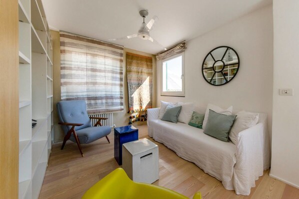 A living room with a white sofa, a blue armchair, a yellow chair, a ceiling fan, and a large window with curtains. - JOIVY il Cielo di Laura (Genoa)