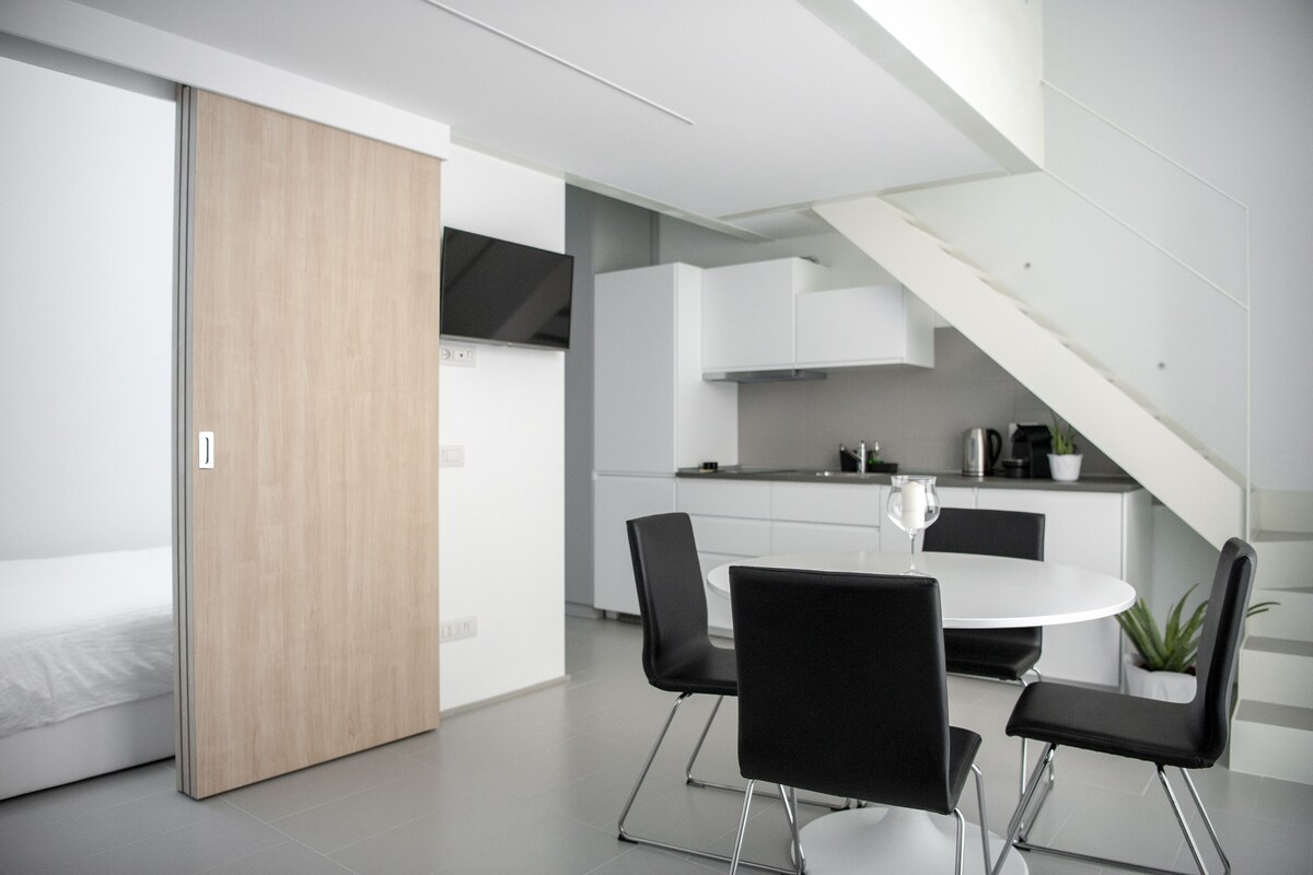 Een moderne keuken met witte kasten, een zwart aanrecht en een eethoek met een witte tafel en zwarte stoelen.