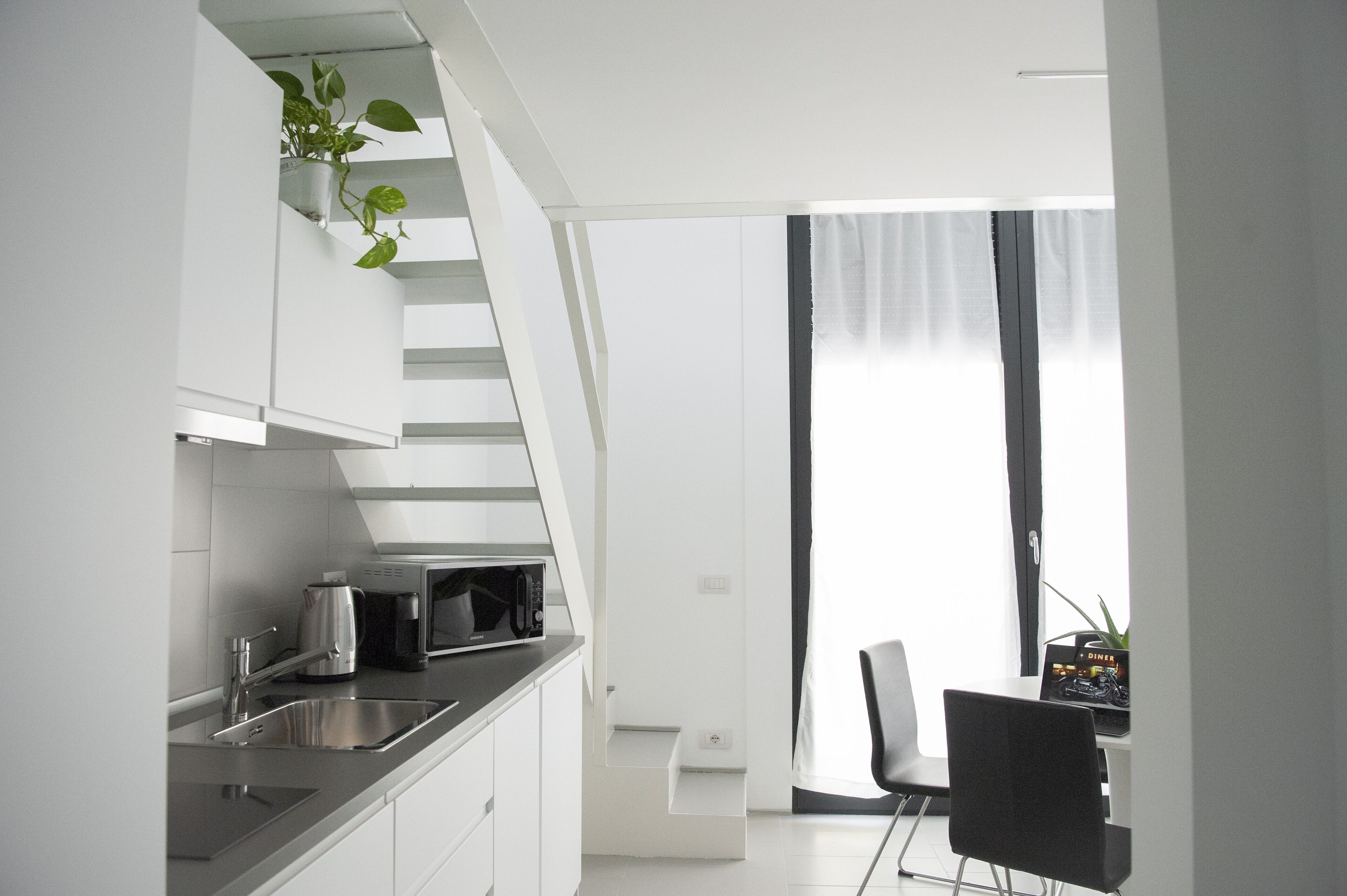 Una cocina moderna con armarios blancos, un lavamanos de acero inoxidable, una cafetera y un microondas. Se ve una escalera que lleva a un nivel superior, y un comedor con una silla y una mesa pequeña.