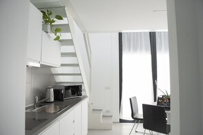 Een moderne keuken met witte kastjes, een roestvrijstalen spoelbak, een koffiezetapparaat en een magnetron. Er is een trap die naar een hoger gelegen verdieping leidt, en een eethoek met een stoel en een kleine tafel.