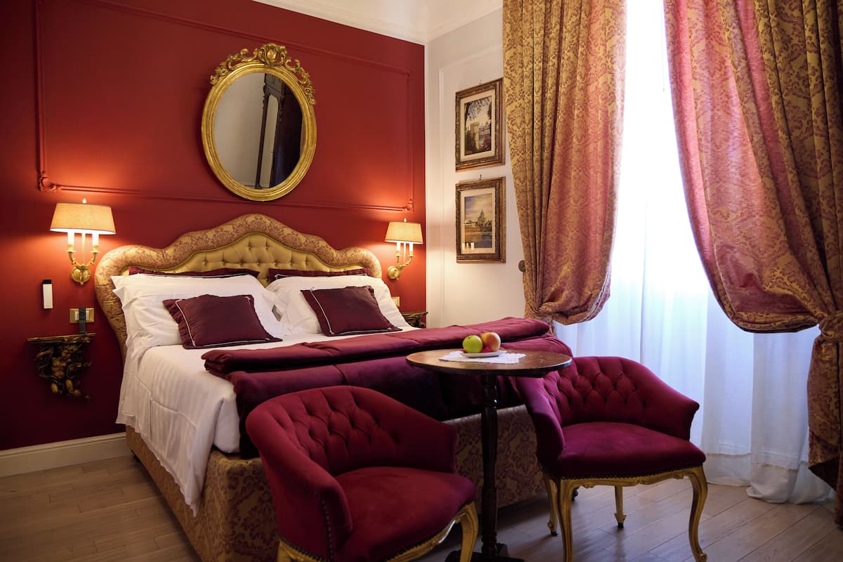 A hotel room with a large bed, two armchairs, a small table with apples, and a mirror on the wall.