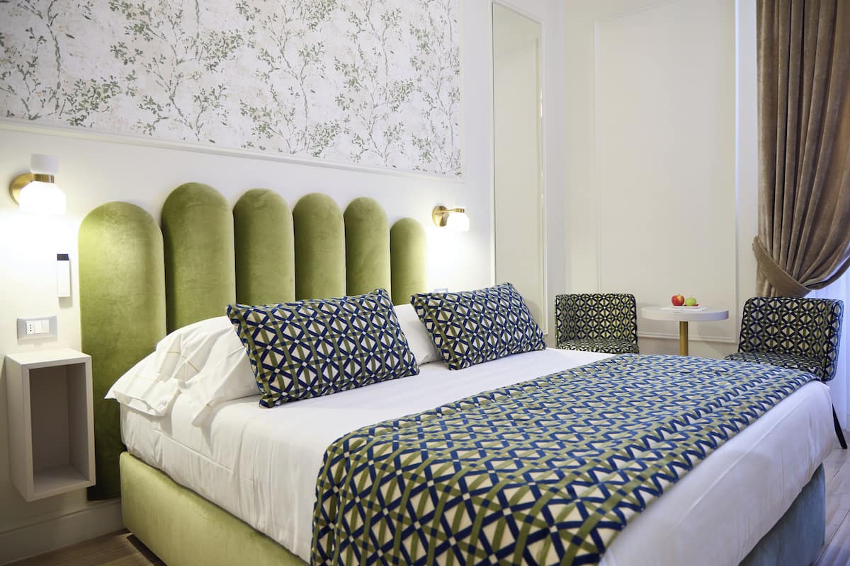 A neatly arranged hotel room with a large bed, green upholstered headboard, patterned pillows, and a small table with fruit.