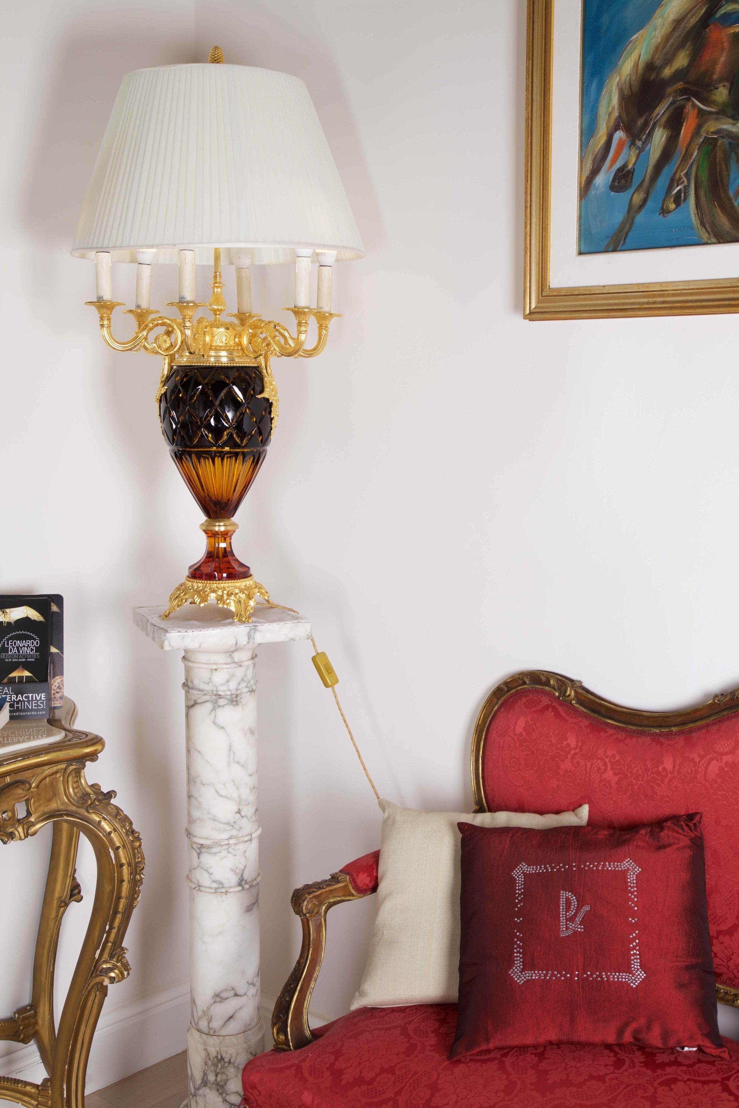Una habitación con suelo de mármol, una butaca tapizada en rojo, una mesa con marco dorado, una lámpara blanca de base dorada y un cuadro enmarcado en la pared.