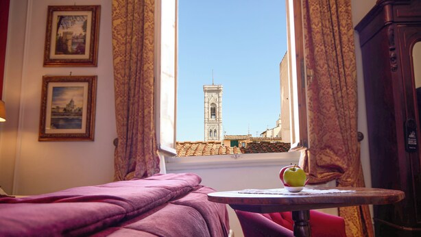 A room with a bed, a table with an apple, and a window with a view of a tower.
