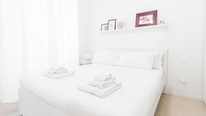 Une chambre à coucher blanche, avec un lit, deux tables de chevet, une étagère murale ornée de tableaux encadrés et une table de chevet avec une lampe.