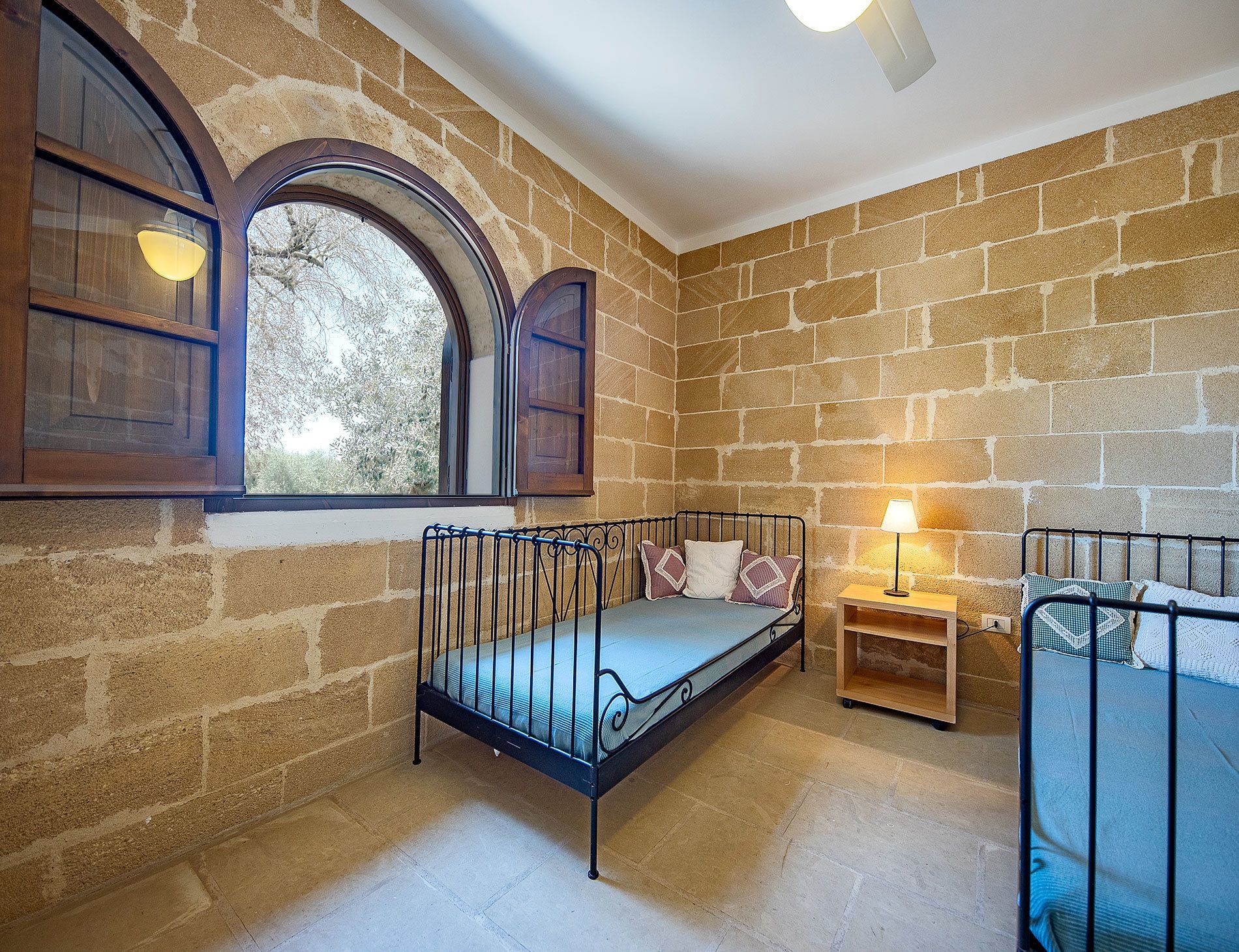 A cozy room with a stone wall, a window with wooden shutters, a metal bed frame, and a bedside table with a lamp.