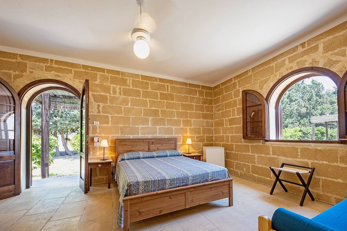 Ein Schlafzimmer mit Steinwand, einem hölzernen Bett, einem Nachttisch mit Lampe, einem kleinen Tisch und einem Fenster mit Blick auf Grünflächen.