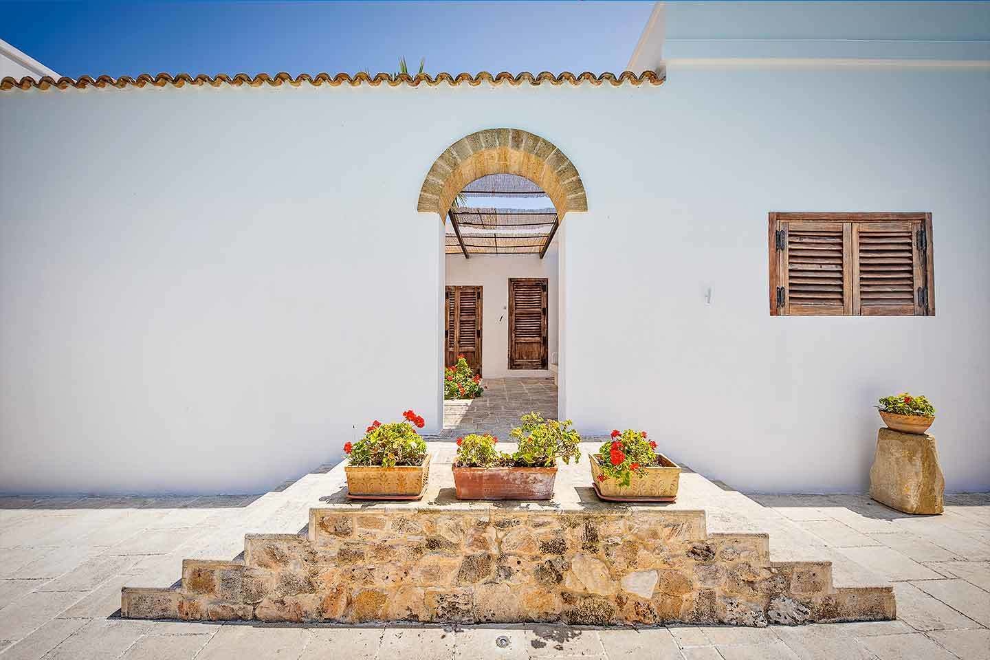 A white building with a stone staircase leading to a wooden door. There are potted plants on the steps and a window with wooden shutters.