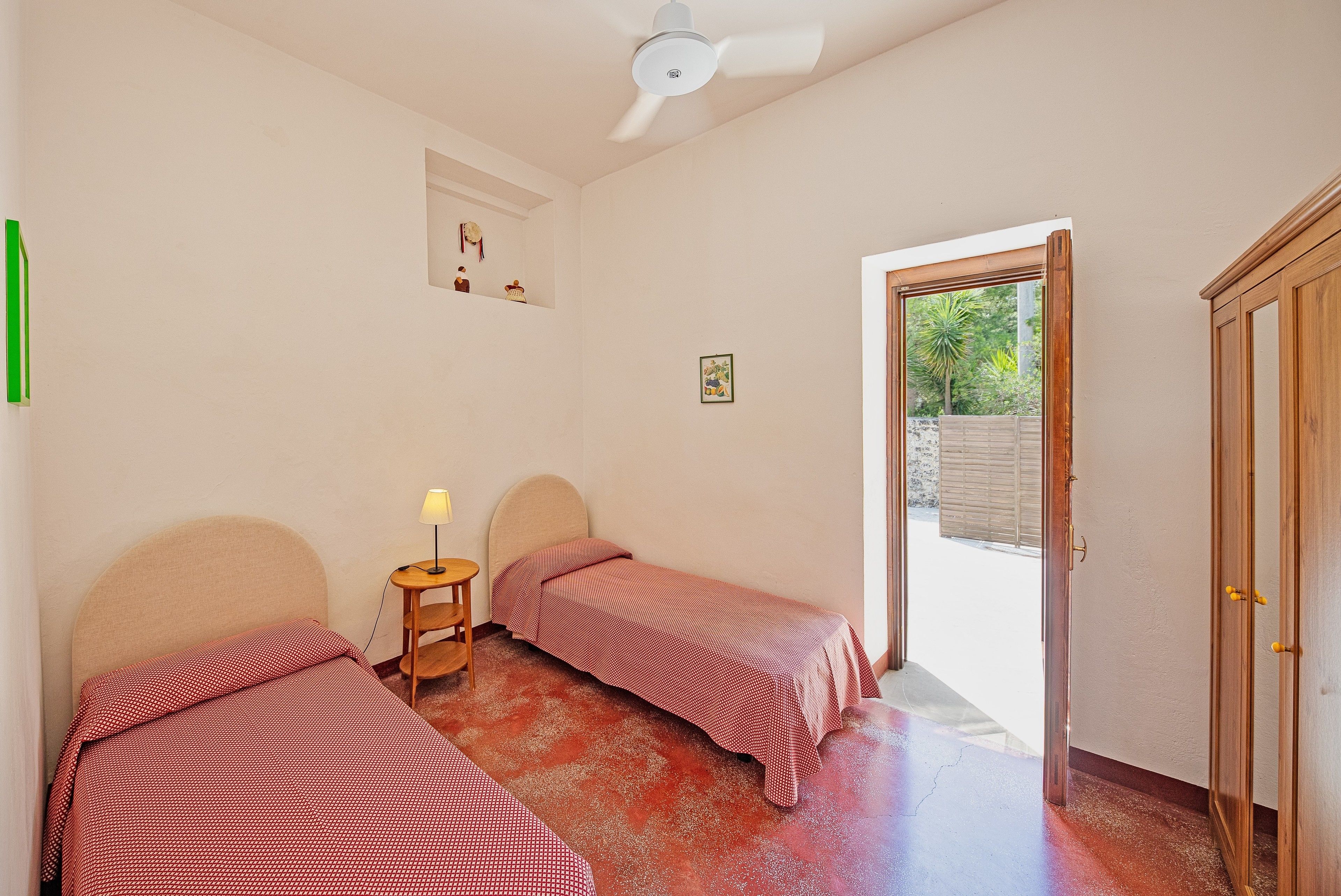 Quarto com duas camas de solteiro, um ventilador de teto, uma mesinha com abajur e uma porta que dá para área externa.