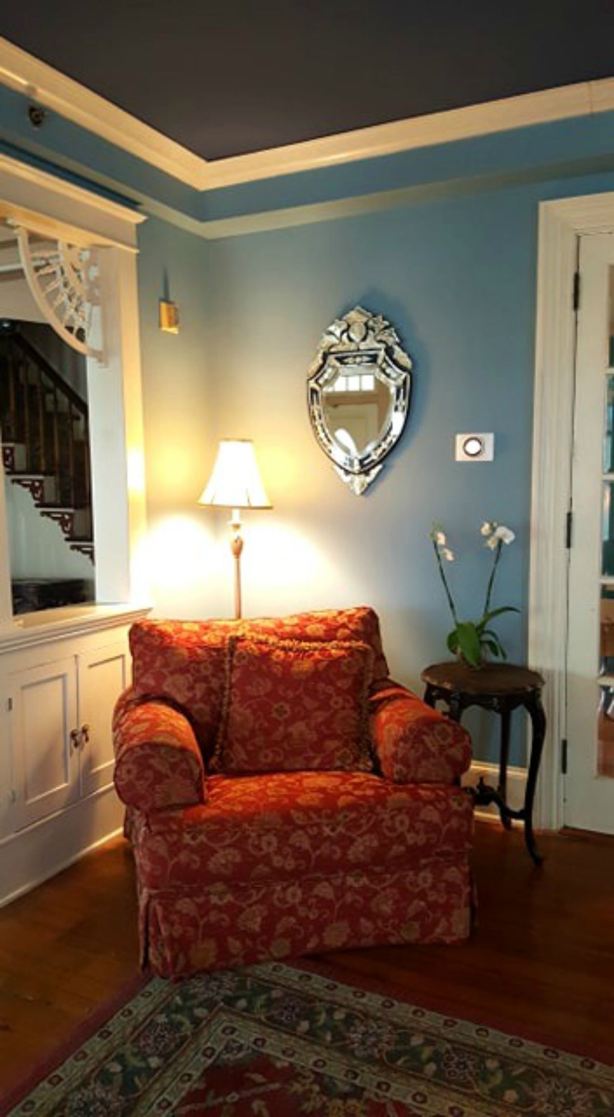 A room with a patterned armchair, a lamp, a mirror, and a staircase.