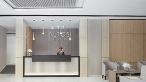Un área de recepción moderna de hotel, con un mostrador de recepción, un hombre de pie detrás de él, y una zona de estar con dos sofás y una mesa de centro.