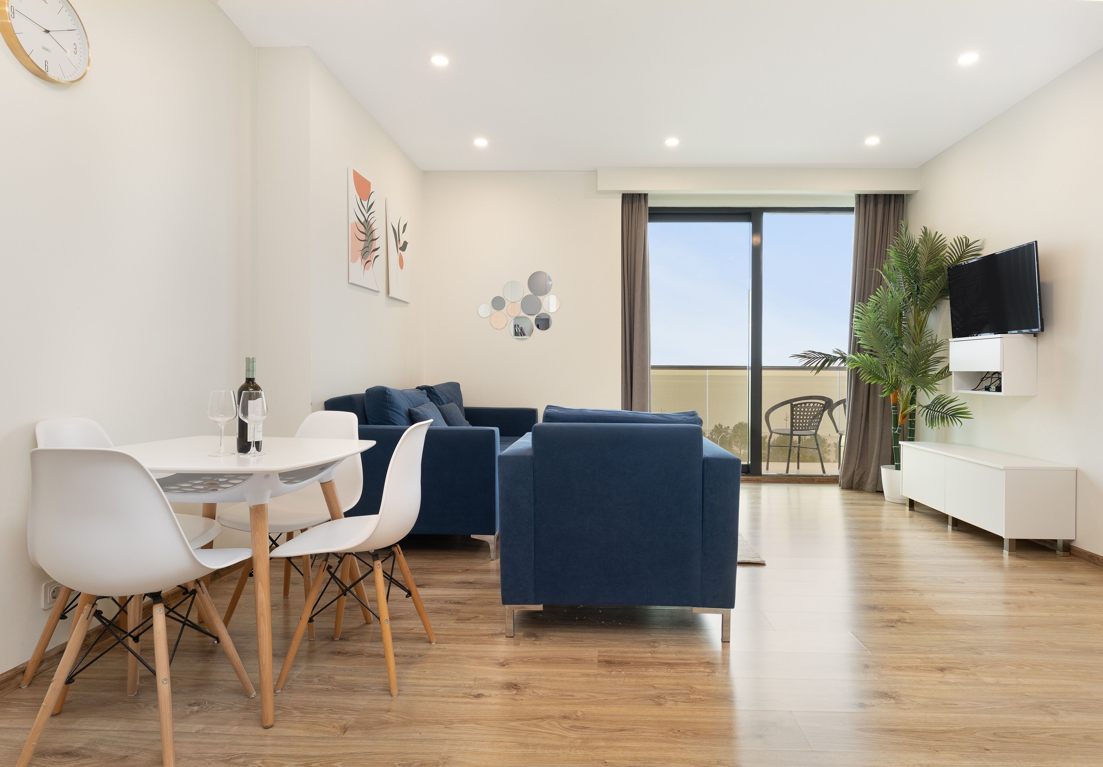 A modern living room with a dining area, a sofa set, a TV, and a balcony with a view.