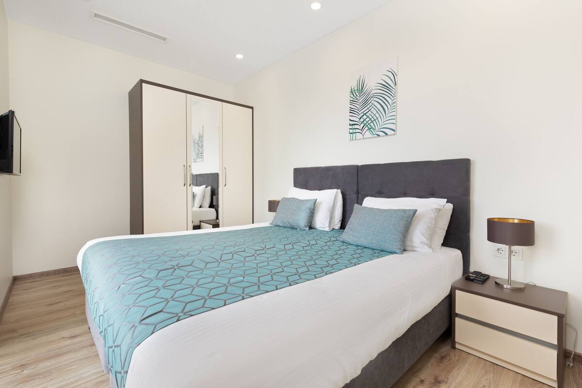 A modern hotel room with a large bed, a bedside table with a lamp, a wardrobe, and a wall-mounted TV.